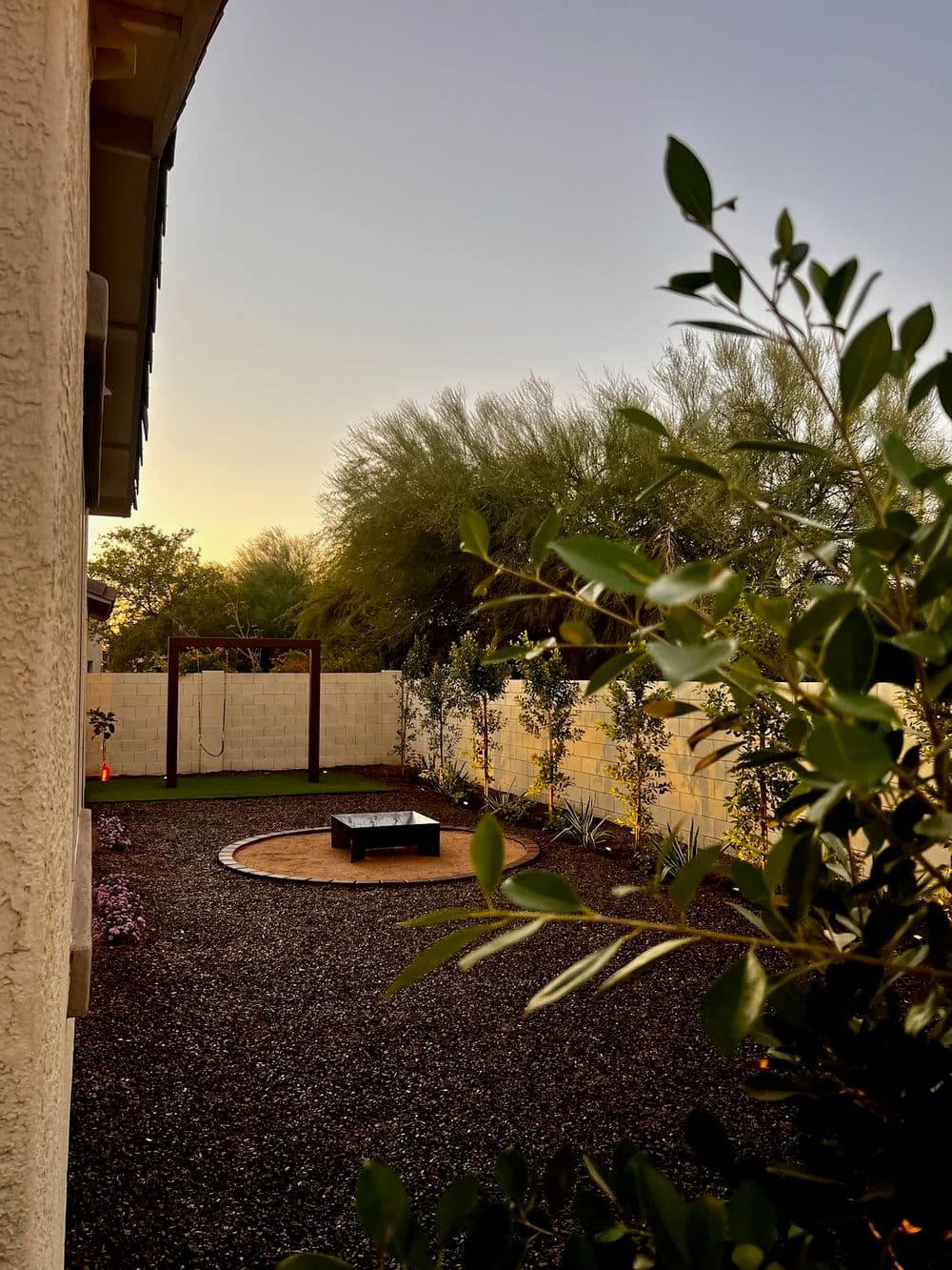 Backyard view featuring a fire pit, swing set, and desert landscaping at sunset.
