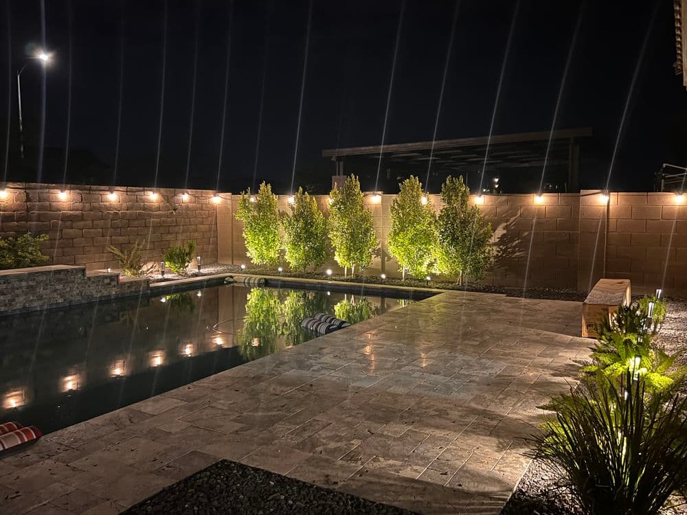 Serene backyard pool at night, illuminated by lights and surrounded by lush greenery.