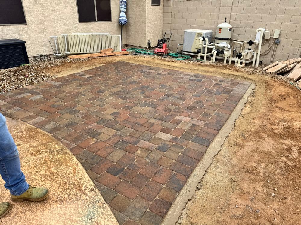 Paver stone patio installation in backyard with utility equipment and landscaping tools nearby.