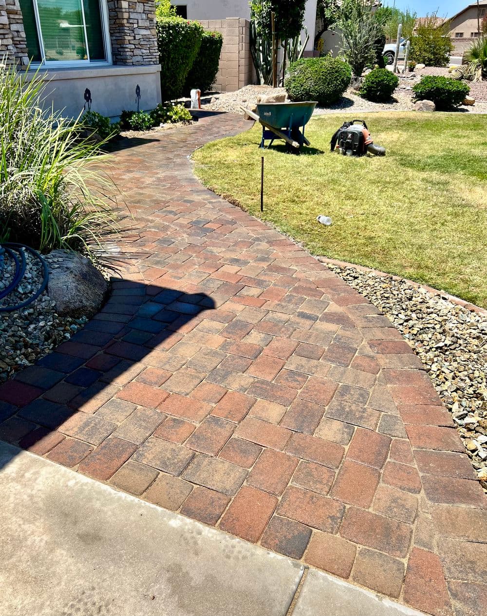 Paver stone walkway in landscaped yard with wheelbarrow and tools nearby.
